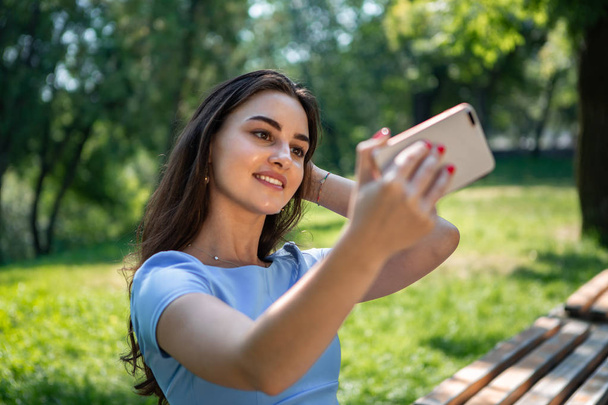 Mosolygó fiatal nő egy kék ruhában ül egy parkban, és így selfie a smartphone. - Fotó, kép