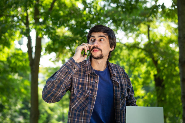 Young brunet freelancer with beard and mustache sitting with a laptop in a park and speaks on the smartphone. Work at any place - Фото, зображення