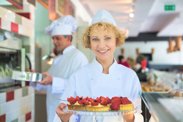 Portrait of smiling mature chef holding fresh strawberry tarts in tray at restaurant - Фото, изображение