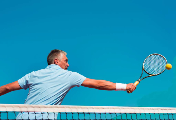 Low angle view of confident mature man hitting tennis ball with racket on court against clear blue sky - Fotó, kép