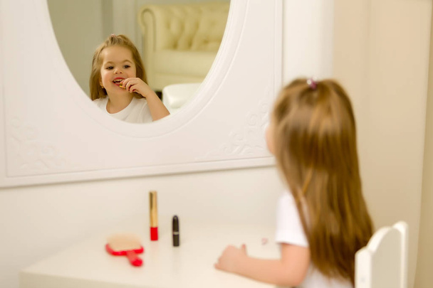 A little girl near the mirror paints lipstick lips - Фото, изображение