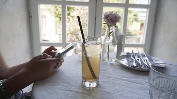 Una joven usa un teléfono inteligente en un café, junto a su refresco de saúco
. - Imágenes, Vídeo
