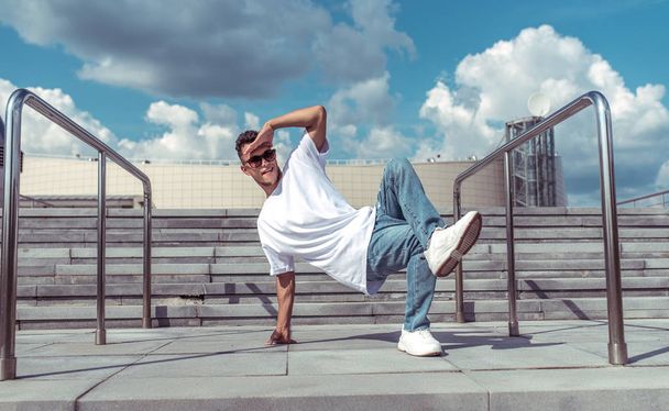 young guy a dancer in a white T-shirt, jeans, dancing break dance in summer in the city, in sunglasses, white sneakers background building clouds, active hip hop, youth lifestyle - Φωτογραφία, εικόνα