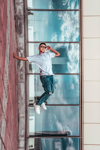 young guy a dancer in a white T-shirt, jeans, dancing break dance in summer in the city, in sunglasses, white sneakers background building clouds, active hip hop, youth lifestyle - Фото, изображение