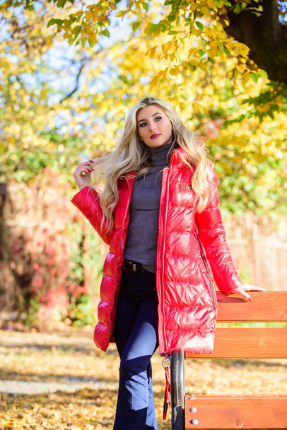 Lady attractive posing in jacket near bench. Woman fashionable blonde with makeup stand in autumnal park. Jacket for fall season concept. Girl wear red bright warm jacket. Fall fashion concept - Photo, Image