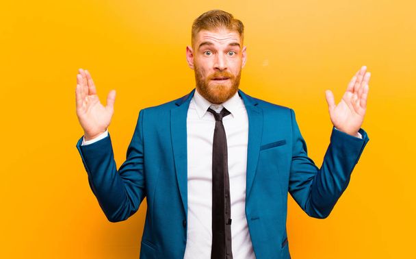 young red head businessman looking shocked and astonished, with jaw dropped in surprise when realizing something unbelievable against orange background - Foto, afbeelding