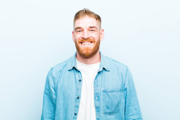 young red head man looking happy and goofy with a broad, fun, loony smile and eyes wide open against soft blue background - Фото, изображение