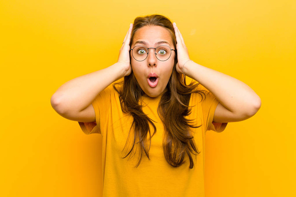 young pretty woman looking unpleasantly shocked, scared or worried, mouth wide open and covering both ears with hands against orange background - Foto, imagen