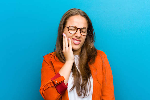 young woman holding cheek and suffering painful toothache, feeling ill, miserable and unhappy, looking for a dentist against blue background - Fotoğraf, Görsel