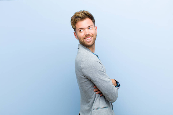 young businessman smiling gleefully, feeling happy, satisfied and relaxed, with crossed arms and looking to the side against blue background - Fotoğraf, Görsel