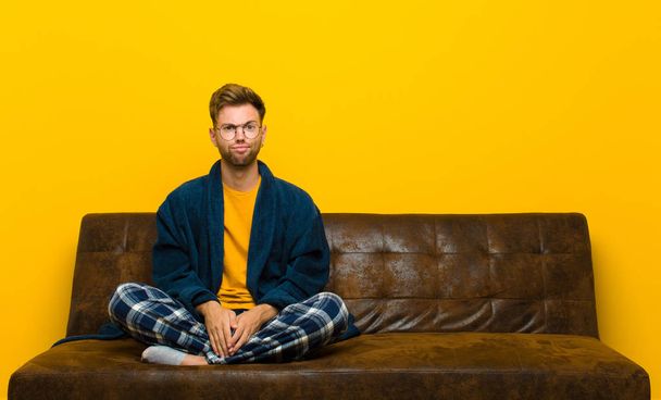 young man wearing pajamas feeling confused and doubtful, wondering or trying to choose or make a decision . sitting on a sofa - Photo, Image
