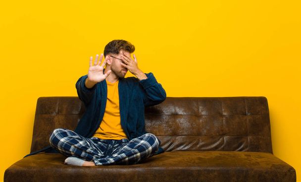young man wearing pajamas covering face with hand and putting other hand up front to stop camera, refusing photos or pictures . sitting on a sofa - 写真・画像