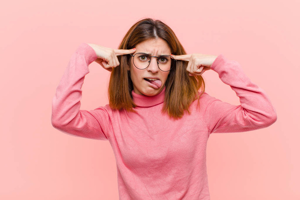 young pretty woman shrugging with a dumb, crazy, confused, puzzled expression, feeling annoyed and clueless against pink background - Photo, Image