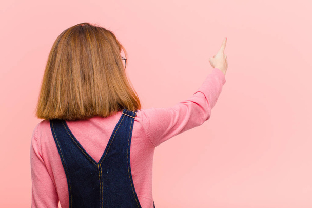 young pretty woman standing and pointing to object on copy space, rear view against pink background - Photo, image