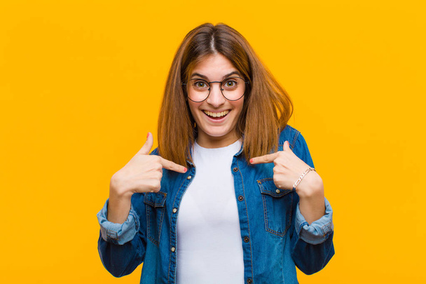 young pretty woman feeling happy, surprised and proud, pointing to self with an excited, amazed look against yellow background - Foto, Imagen