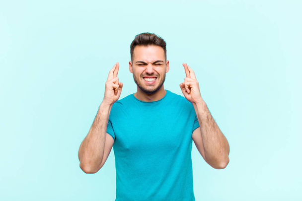 young hispanic man smiling and anxiously crossing both fingers, feeling worried and wishing or hoping for good luck against blue background - Photo, image