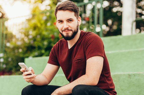 Attractive young smiling man using phone in a public park - Zdjęcie, obraz