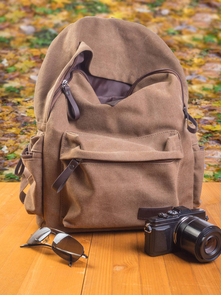 Туристический рюкзак, фотоаппарат и очки на фоне осенних листьев
 - Фото, изображение