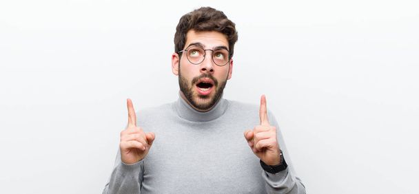 young manager man looking shocked, amazed and open mouthed, pointing upwards with both hands to copy space against white wall - Фото, изображение