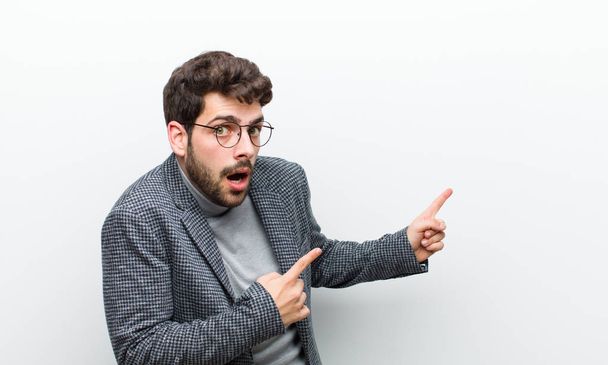 young manager man feeling shocked and surprised, pointing to copy space on the side with amazed, open-mouthed look against white wall - Foto, Bild