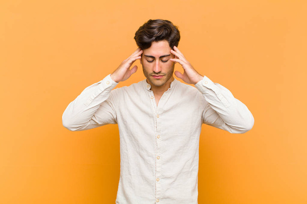 young handsome man looking concentrated, thoughtful and inspired, brainstorming and imagining with hands on forehead against orange background - Foto, Bild