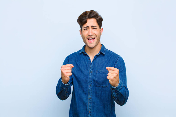 young handsome man feeling shocked, excited and happy, laughing and celebrating success, saying wow! against blue background - Фото, зображення