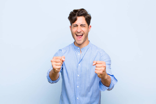 young handsome man feeling happy, surprised and proud, shouting and celebrating success with a big smile against blue background - Фото, зображення