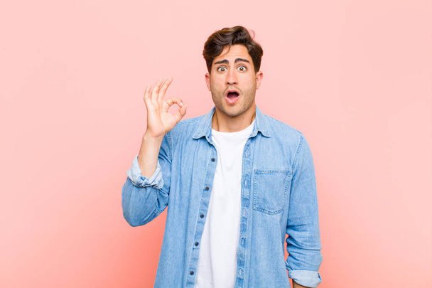 young handsome man feeling successful and satisfied, smiling with mouth wide open, making okay sign with hand against pink background - Fotografie, Obrázek