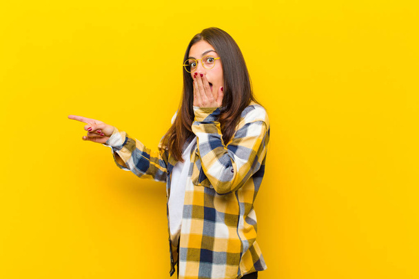 young  pretty woman feeling happy, shocked and surprised, covering mouth with hand and pointing to lateral copy space against orange wall - 写真・画像