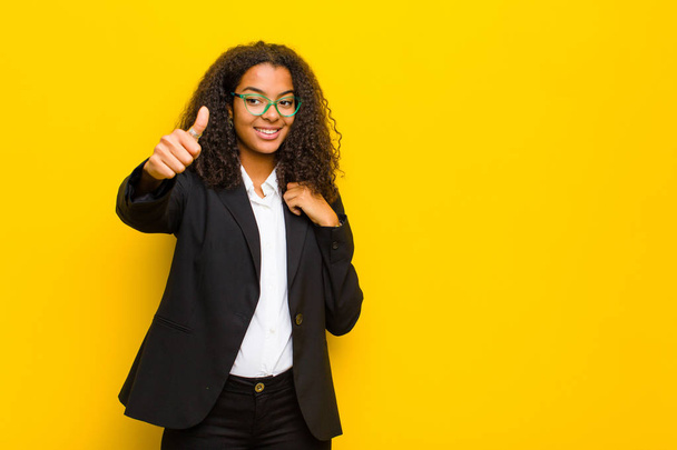 black business woman feeling proud, carefree, confident and happy, smiling positively with thumbs up against orange wall - Photo, Image