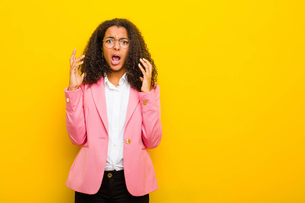 black business woman screaming with hands up in the air, feeling furious, frustrated, stressed and upset against orange wall - Фото, изображение