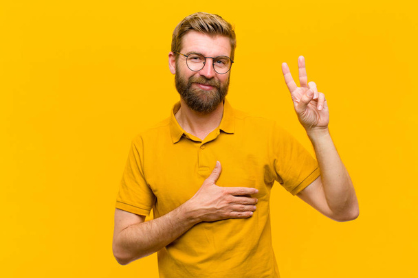 young blonde man looking happy, confident and trustworthy, smiling and showing victory sign, with a positive attitude against orange wall - Foto, Imagen