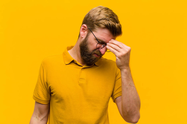 young blonde man feeling stressed, unhappy and frustrated, touching forehead and suffering migraine of severe headache against orange wall - Photo, Image