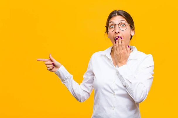 young businesswoman feeling happy, shocked and surprised, covering mouth with hand and pointing to lateral copy space against orange wall - Fotó, kép