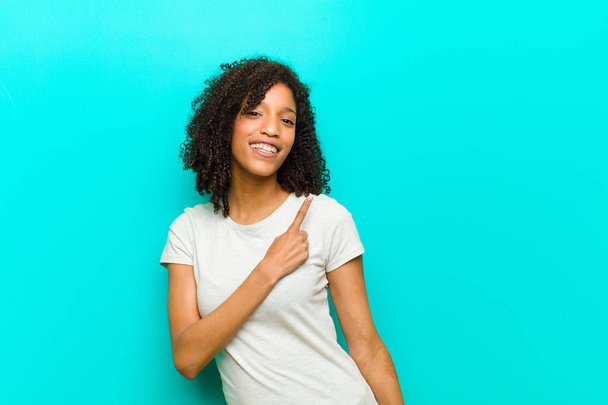 joven mujer negra sonriendo alegremente, sintiéndose feliz y señalando hacia un lado y hacia arriba, mostrando el objeto en el espacio de copia contra la pared azul
 - Foto, Imagen