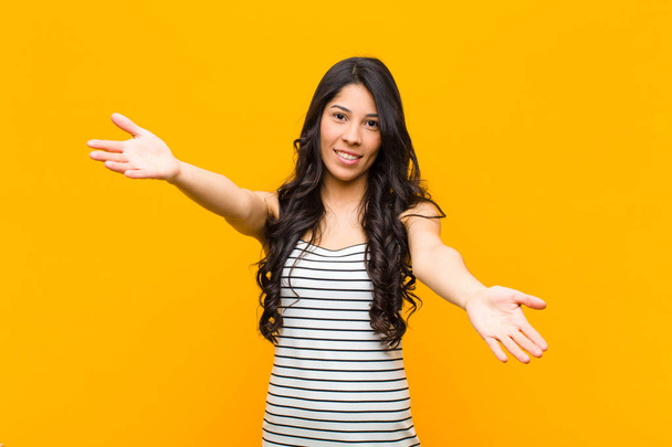 young pretty latin woman smiling cheerfully giving a warm, friendly, loving welcome hug, feeling happy and adorable against orange wall - Photo, Image