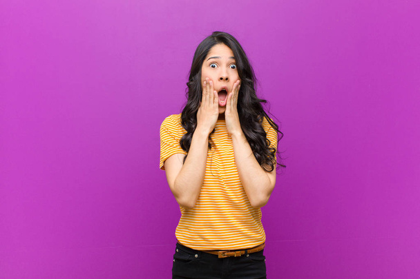 young pretty latin woman feeling shocked and scared, looking terrified with open mouth and hands on cheeks against purple wall - Фото, изображение