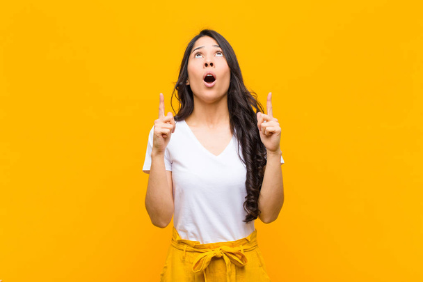 young pretty latin woman looking shocked, amazed and open mouthed, pointing upwards with both hands to copy space against orange wall - Photo, image