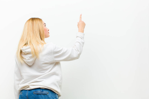 young blonde woman standing and pointing to object on copy space, rear view against white wall - Valokuva, kuva