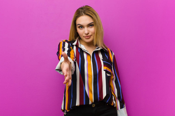 young blonde woman smiling, looking happy, confident and friendly, offering a handshake to close a deal, cooperating against purple wall - Foto, Bild
