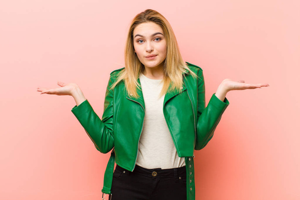 young pretty blonde woman feeling puzzled and confused, doubting, weighting or choosing different options with funny expression against pink flat wall - Foto, Imagen