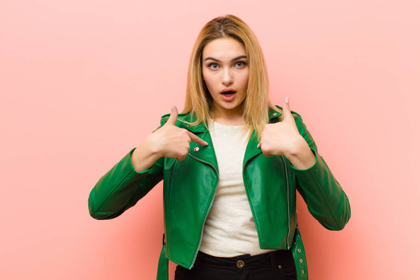 young pretty blonde woman feeling happy, surprised and proud, pointing to self with an excited, amazed look against pink flat wall - Фото, зображення