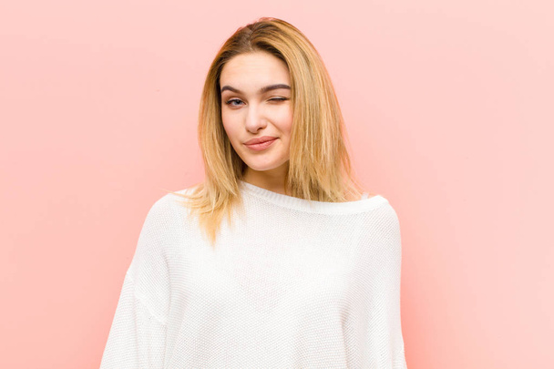 young pretty blonde woman looking happy and friendly, smiling and winking an eye at you with a positive attitude against pink flat wall - Zdjęcie, obraz