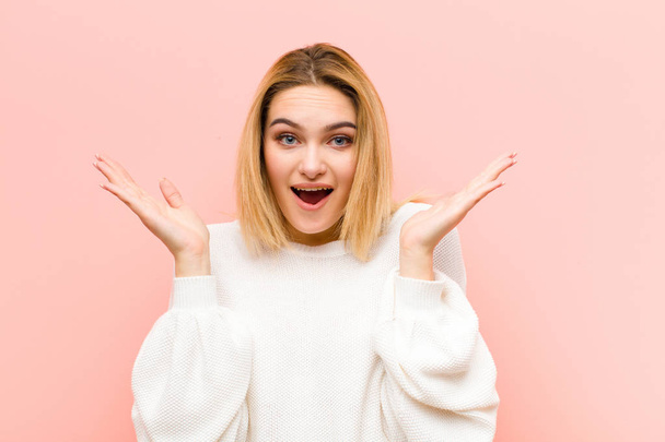 young pretty blonde woman looking happy and excited, shocked with an unexpected surprise with both hands open next to face against pink flat wall - Photo, image