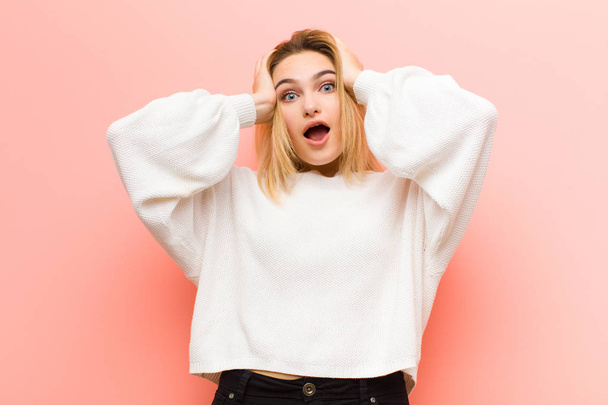 young pretty blonde woman raising hands to head, open-mouthed, feeling extremely lucky, surprised, excited and happy against pink flat wall - Photo, image