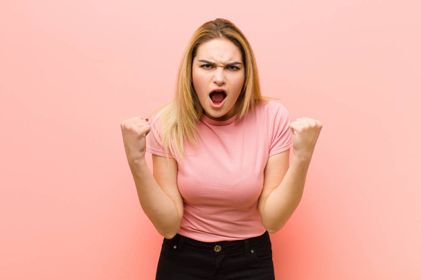 young pretty blonde woman shouting aggressively with an angry expression or with fists clenched celebrating success against pink flat wall - Foto, Imagem