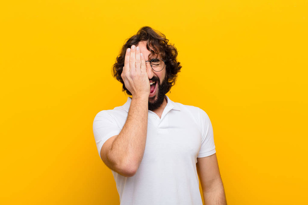 young crazy man looking sleepy, bored and yawning, with a headache and one hand covering half the face against yellow wall - Fotoğraf, Görsel