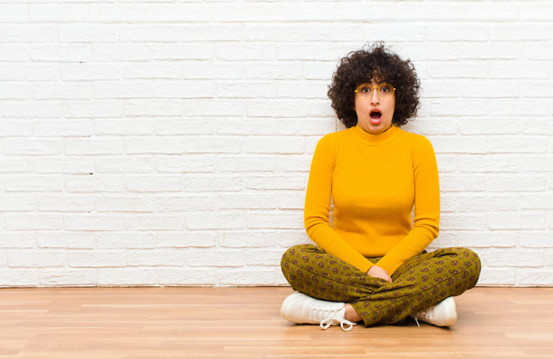 young pretty afro woman feeling terrified and shocked, with mouth wide open in surprise sitting on the floor - Photo, Image