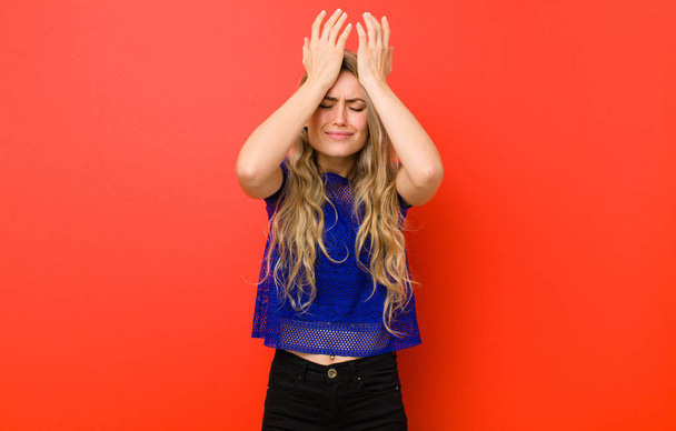 young blonde woman feeling stressed and anxious, depressed and frustrated with a headache, raising both hands to head against red wall - Fotoğraf, Görsel