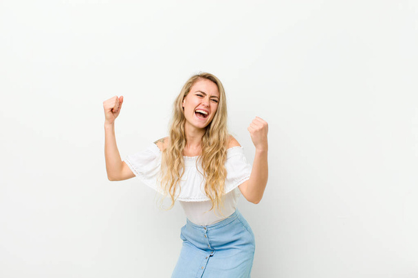 young blonde woman shouting triumphantly, looking like excited, happy and surprised winner, celebrating against white wall - Foto, afbeelding